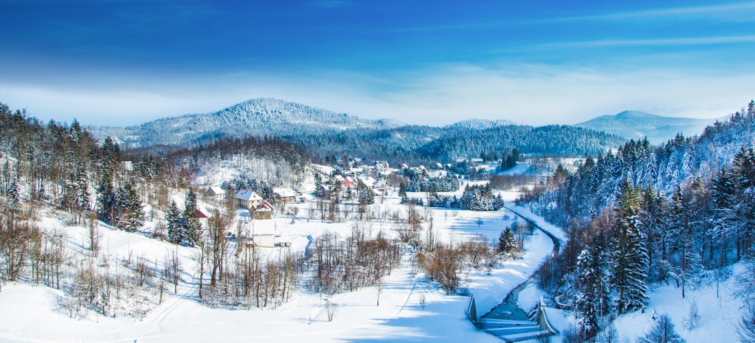 Winterliche Landschaft mit schneebedeckten Hügeln, einem kleinen Dorf mit verstreuten Häusern und einer von Schnee gesäumten Straße, die sich durch das Tal schlängelt. Im Hintergrund erstrecken sich bewaldete Berge, deren Bäume ebenfalls mit Schnee bedeck