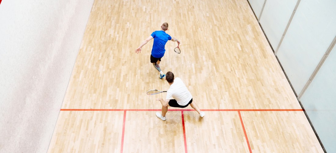Zwei Männer spielen Squash auf einem hellen Holzplatz. Der eine trägt ein blaues Shirt und schlägt den Ball, während der andere in einem weißen Shirt bereitsteht. Das Spielfeld ist von roten Linien begrenzt.