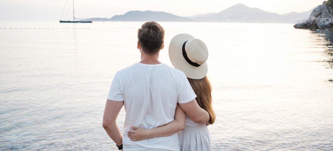 Ein verliebtes Paar steht Arm in Arm am Strand und blickt auf das Meer bei Sonnenuntergang. Im Hintergrund ist ein Segelboot zu sehen