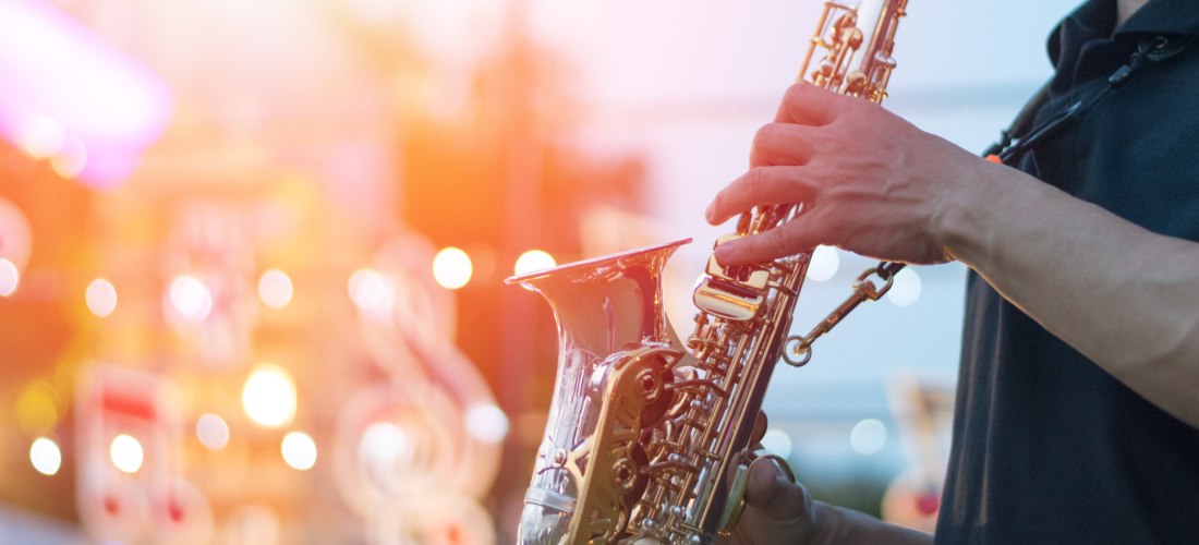 Das Bild zeigt einen Musiker, der ein glänzendes Saxofon spielt. Die Szene ist stimmungsvoll beleuchtet, mit warmem Licht und verschwommenen Musiknoten im Hintergrund – es wirkt wie ein Konzert oder ein Musikfestival bei Sonnenuntergang.