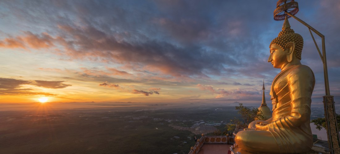 Tiger Cave Temple in Krabi, Thailand: goldene Buddha-Statue bei Sonnenaufgang mit weitem Blick über die Landschaft.