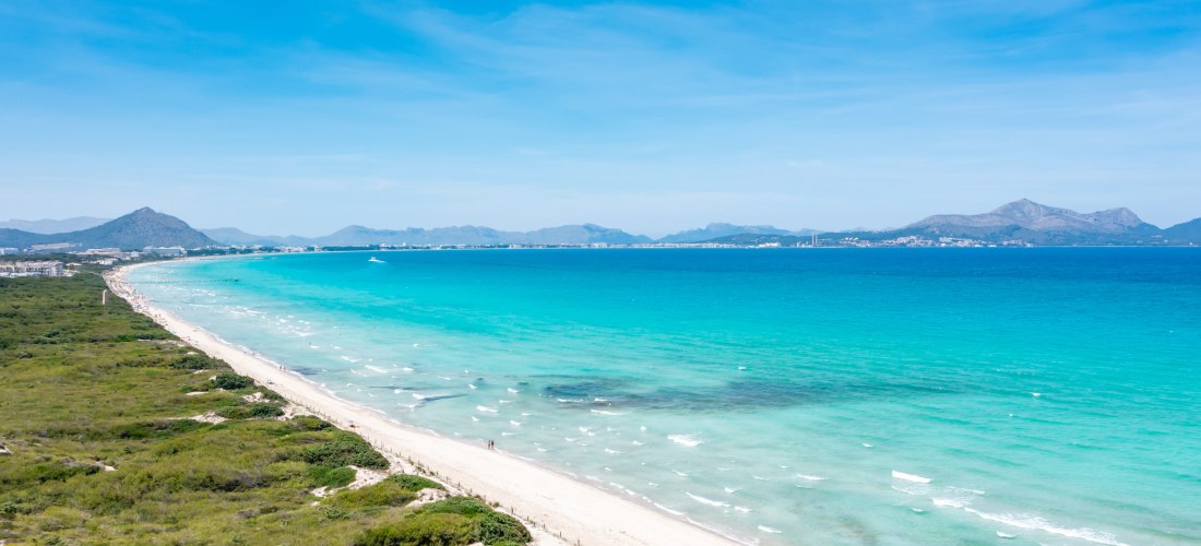Panoramablick über einen langen, weißen Sandstrand mit türkisblauem Meer. Links grüne Dünenlandschaft, rechts die Bucht mit sanften Wellen, im Hintergrund Berge und ferne Küstenlinie