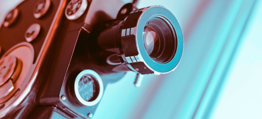 Nahaufnahme einer analogen Filmkamera mit großem Objektiv, warm-kalt ausgeleuchtet. Die Kamera ist auf einen hellen Hintergrund gerichtet und vermittelt Retro-Filmästhetik.