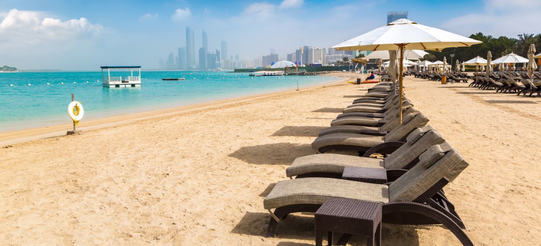 Luxuriöser Sandstrand in Abu Dhabi mit Liegestühlen und Sonnenschirmen, mit Blick auf die Skyline der Stadt im Hintergrund