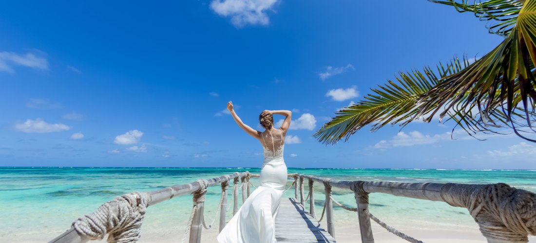 Frau in weißem Kleid steht auf einem Holzsteg am tropischen Strand und blickt auf das türkisfarbene Meer unter blauem Himmel