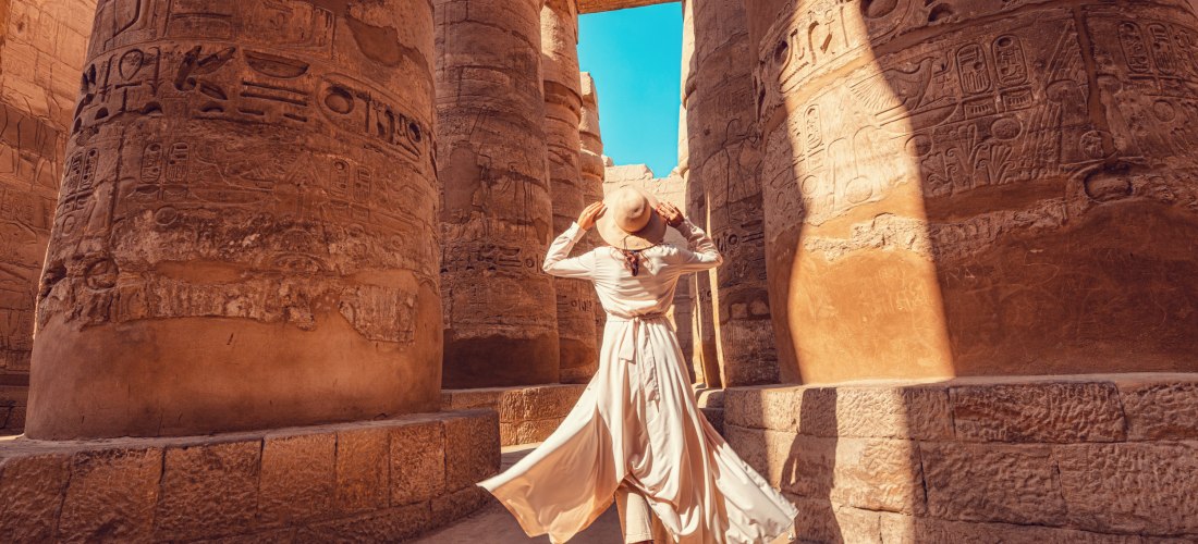 Eine Frau in einem langen, hellen Kleid und Hut steht zwischen monumentalen Säulen eines antiken ägyptischen Tempels. Die Säulen sind mit Hieroglyphen verziert. Die Frau blickt nach oben, während Sonnenlicht und blauer Himmel die Szene erhellen.