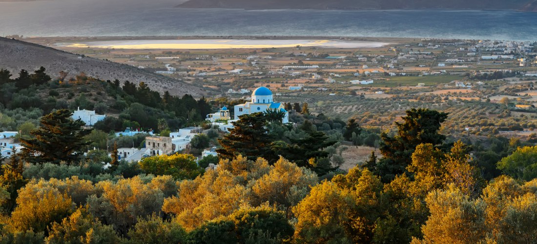 Malerischer Sonnenuntergang über griechischem Dorf mit weißer Kirche und blauer Kuppel, umgeben von grüner Hügellandschaft, Olivenhainen und dem Meer in der Ferne.