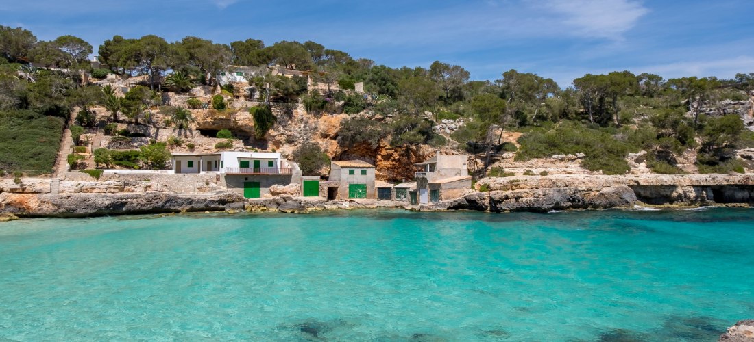 Traditionelle Bootshäuser mit grünen Türen am türkisfarbenen Wasser der Bucht Cala Llombards auf Mallorca, umgeben von Felsen und Pinienbäumen