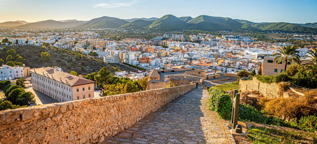 Steinerner Weg auf der Festungsmauer Dalt Vila mit weitem Blick über Ibiza-Stadt bis zu grünen Hügeln im Abendlicht.