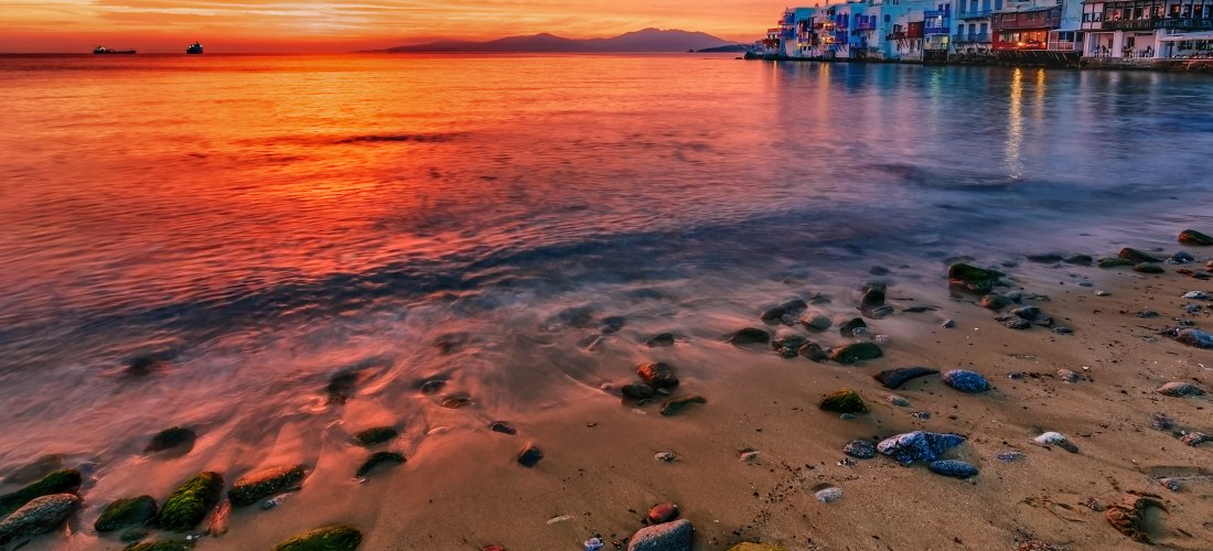 Sonnenuntergang über dem Viertel Little Venice auf Mykonos mit reflektierendem Licht im Meer und bunten Häusern am Ufer