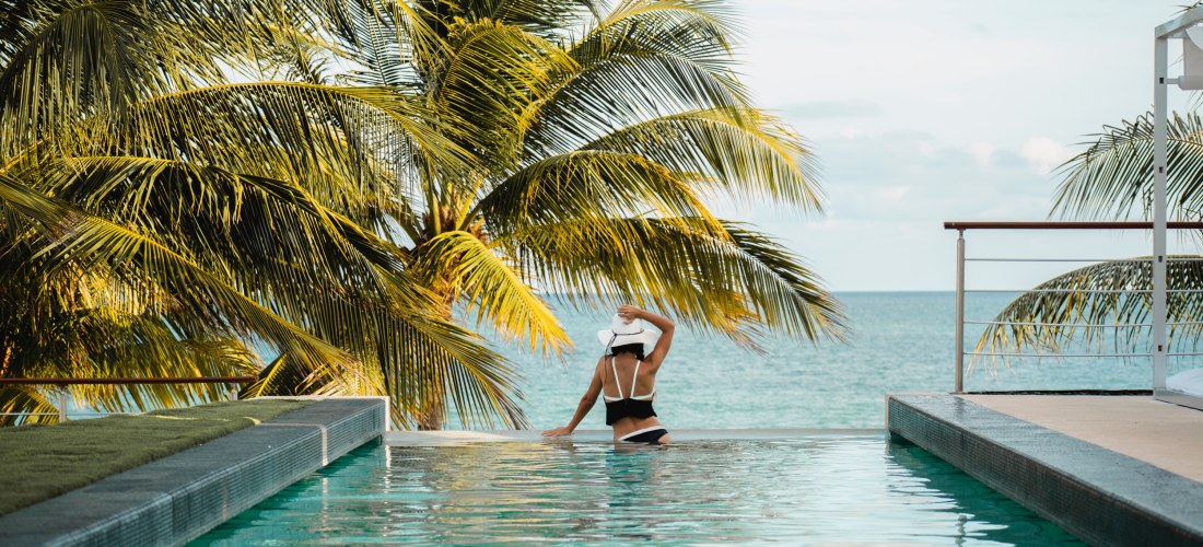 Infinity-Pool mit einer Frau in einem weißen Hut, die auf das Meer blickt, umgeben von Palmen und tropischer Vegetation
