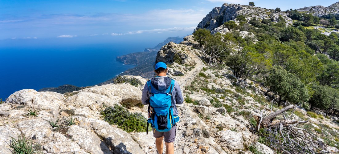 Wanderer mit blauem Rucksack auf einem felsigen Pfad im Tramuntana-Gebirge auf Mallorca, mit Blick auf das Meer und die Küstenlinie