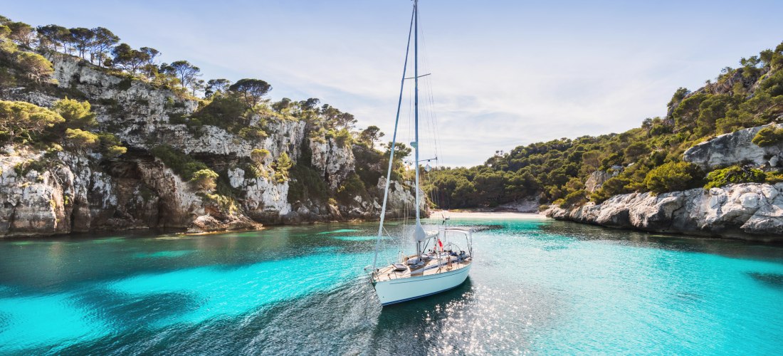 Ein einsames Segelboot gleitet durch kristallklares, türkisfarbenes Wasser in einer malerischen Bucht. Umgeben ist die Szene von felsigen Klippen und üppigem Pinienwald. Die Sonne spiegelt sich glitzernd auf dem Wasser, der Himmel ist strahlend blau – ein