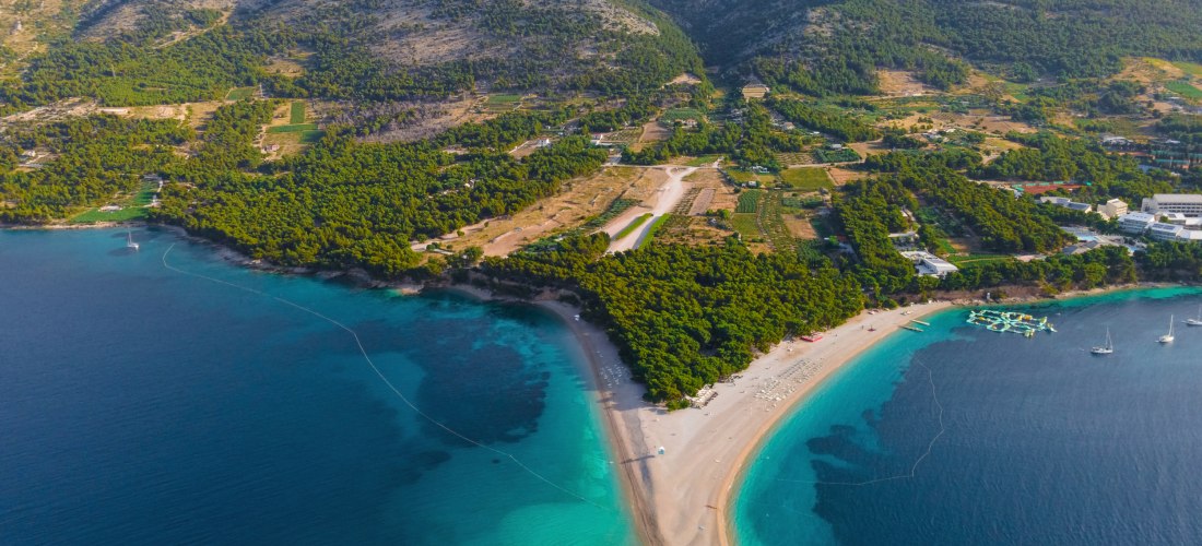 Kroatien - Zlatni Rat Luftaufnahme des bekannten Zlatni Rat Strandes auf der kroatischen Insel Brač. Die landzungeförmige Kiesbank ragt sichelförmig ins türkisfarbene Meer. Umgeben ist der Strand von dichten Pinienwäldern und einer bergigen Küstenlandschaft im Hintergrund. Ein
