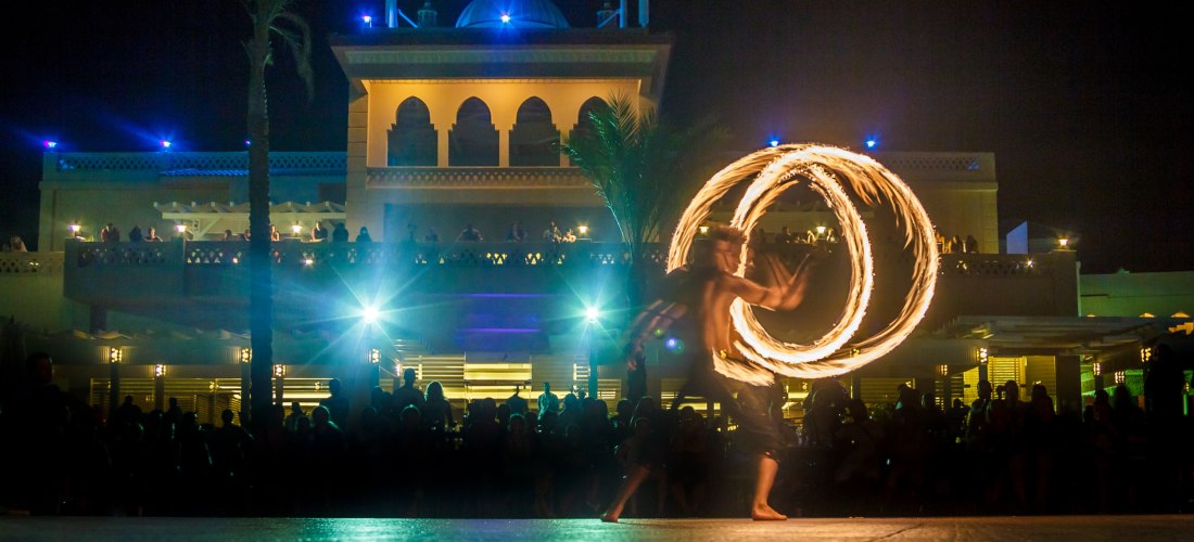 Ein Feuerspucker zeigt bei Nacht eine beeindruckende Feuershow mit zwei brennenden Kreisen vor einem orientalisch anmutenden, hell beleuchteten Gebäude mit Kuppel. Viele Zuschauer beobachten die Performance unter freiem Himmel.
