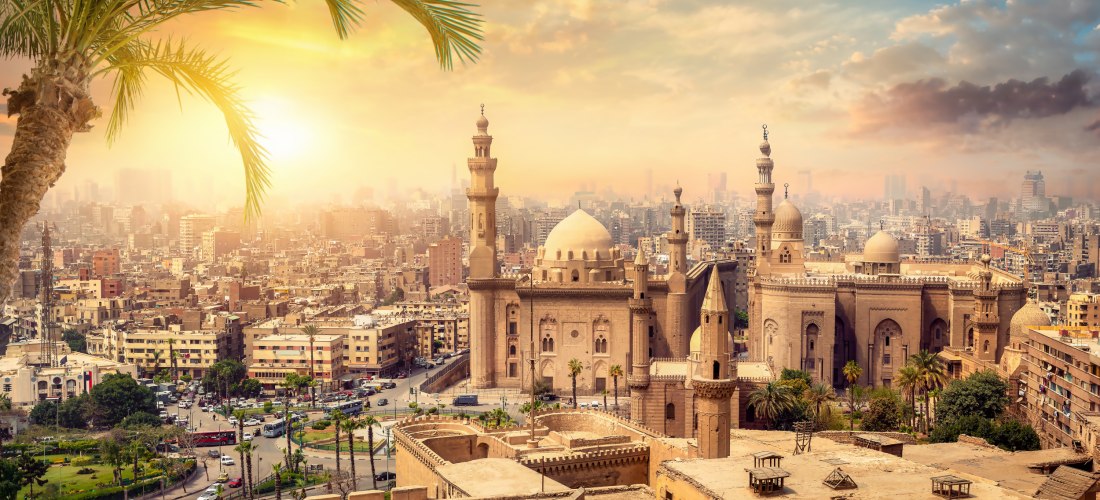 Das Bild zeigt ein weitläufiges Panorama der ägyptischen Hauptstadt Kairo bei Sonnenuntergang. Im Vordergrund steht die eindrucksvolle Sultan-Hassan-Moschee mit ihrer großen Kuppel und mehreren Minaretten, umgeben von Palmen und historischen Mauern. Die m