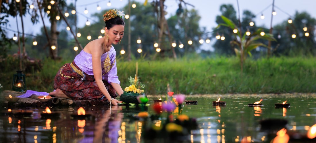 Das Bild zeigt eine Frau in prachtvoller traditioneller thailändischer Kleidung, die in ruhiger Haltung ein mit Blumen und Kerze geschmücktes Krathong auf einem Teich platziert. Es ist Dämmerung, im Hintergrund leuchten warme Lichterketten über der Wiese 