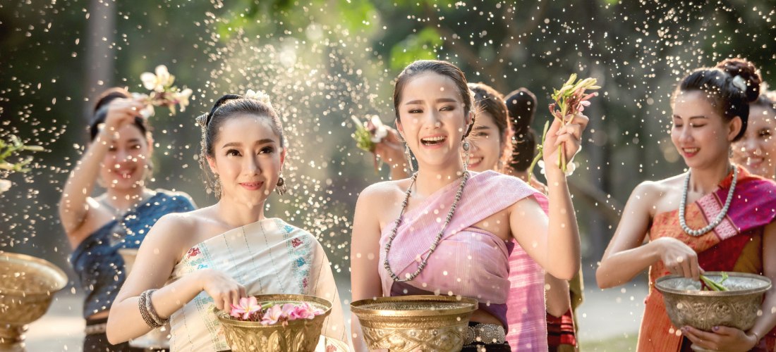 Das Bild zeigt eine fröhliche Gruppe junger Frauen in traditioneller thailändischer Kleidung während einer Feier im Freien. Sie tragen elegante Seidengewänder in kräftigen Farben und halten vergoldete Schalen, gefüllt mit Blumen und Wasser. In der Luft sp