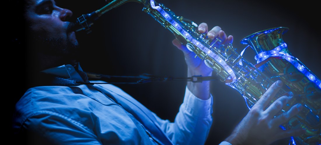 Ein Musiker spielt Saxophon im Dunkeln. Das Instrument ist mit blauen LED-Lichtern beleuchtet. Der Mann trägt ein helles Hemd und ist konzentriert auf sein Spiel. Die Szene wirkt stilvoll und stimmungsvoll.