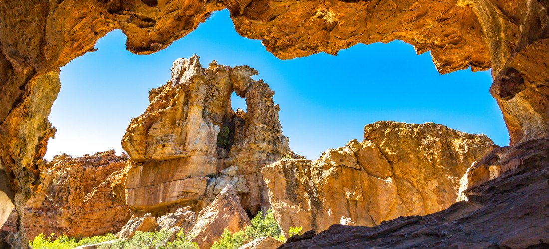 Blick aus einer natürlichen Felsöffnung auf bizarre, rot-orange Felsformationen unter strahlend blauem Himmel. Die Szene zeigt eine beeindruckende Landschaft mit zerklüfteten Gesteinsformationen und grünem Buschwerk im Vordergrund, typisch für die Cederbe