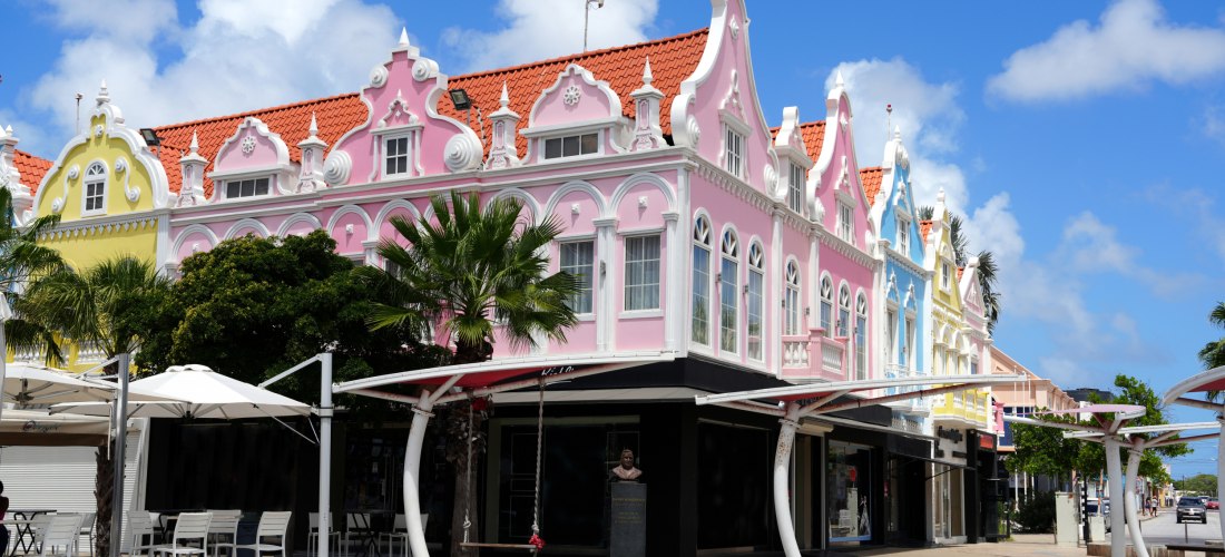 Reihe farbenfroher, kolonial inspirierter Gebäude in Oranjestad, Aruba. Die Fassaden sind in Rosa, Gelb und Blau gestrichen, mit weißen Ornamenten und roten Ziegeldächern. Palmen und weiße Sonnenschirme ergänzen das Straßenbild.