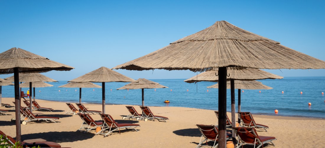 Strand mit Sonnenliegen und Strohdächern vor ruhigem, blauem Meer unter klarem Himmel