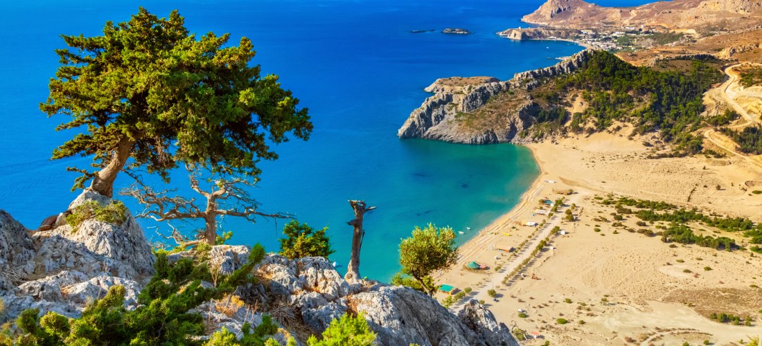 Atemberaubender Blick von einem felsigen Aussichtspunkt mit knorrigen Kiefern auf eine türkisfarbene Bucht. Unten liegt ein breiter Sandstrand mit einzelnen Sonnenschirmen und kleinen Strandhütten. Die Bucht wird eingerahmt von steilen Felsen und Hügeln, 