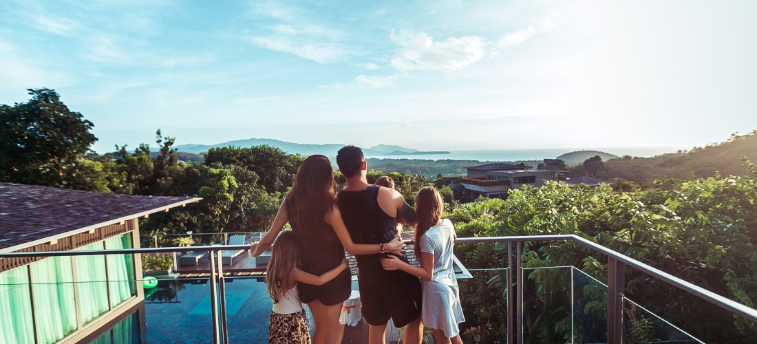 Eine Familie mit zwei Erwachsenen und drei Kindern steht barfuß auf einer modernen Terrasse mit Glasgeländer. Sie schauen gemeinsam in Richtung einer üppig grünen Landschaft mit Bäumen, vereinzelten Gebäuden und einem Blick bis zum Meer oder einem See in 