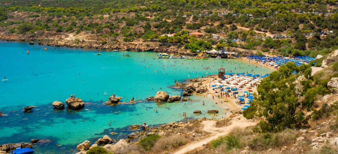 Konnos Bay bei Agia Napa/Protaras: geschützte Sandbucht mit türkisfarbenem Wasser, Felsen und Reihen blauer Sonnenschirme.