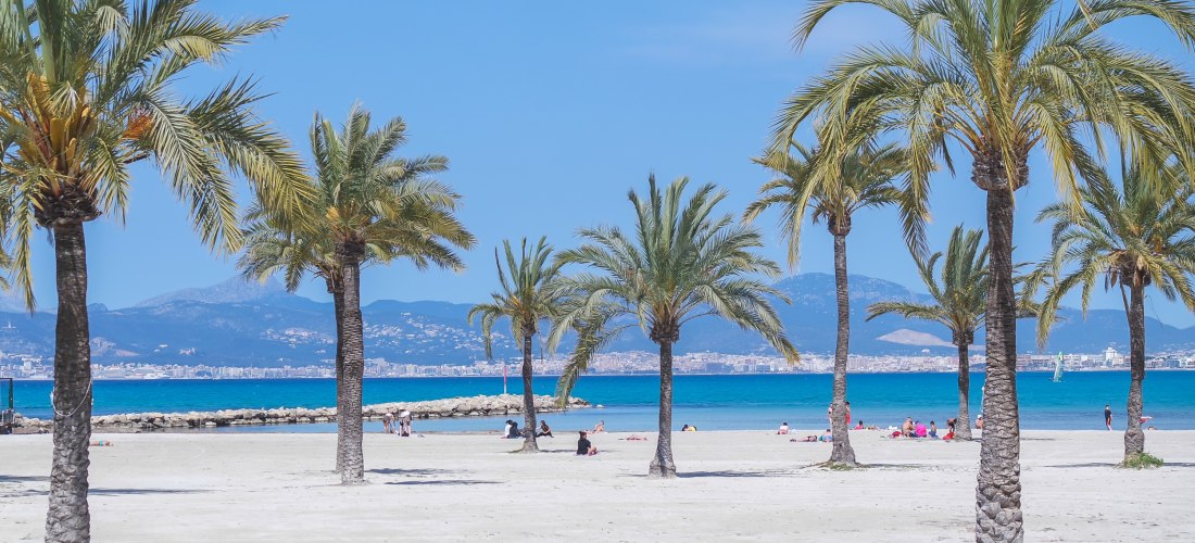 Weißer Sandstrand mit Palmen und Blick auf das türkisfarbene Meer und eine Stadt vor einer Bergkulisse im Hintergrund
