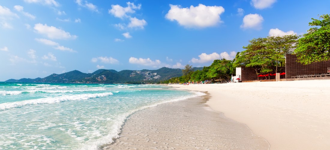 Weißer Sandstrand mit sanften Wellen und türkisblauem Wasser am Chaweng Beach auf Koh Samui, Thailand, mit grünen Bergen im Hintergrund
