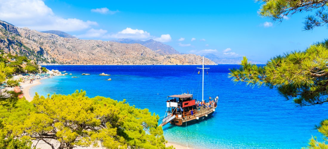 Traumhafte Mittelmeerbucht mit türkisblauem Wasser, Pinienbäumen und einem Holzsegelschiff am weißen Kiesstrand vor einer felsigen Berglandschaft.