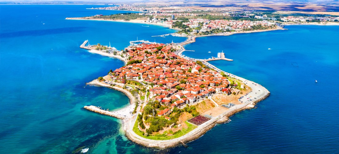 Luftaufnahme einer Halbinsel mit einer historischen Altstadt voller roter Ziegeldächer, umgeben von tiefblauem Meer. Die Stadt ist über eine schmale Landverbindung mit dem Festland verbunden. Im Hintergrund weite Ebenen und weitere Ortschaften.