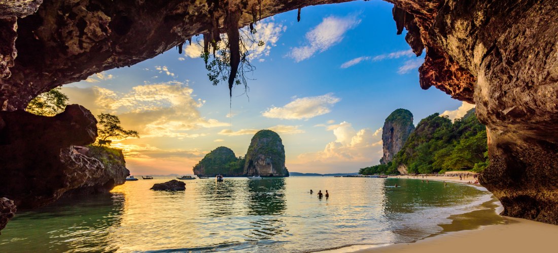 Sonnenuntergang am Phra Nang Cave Beach in Krabi: Blick aus der Höhle auf Meer, Karstfelsen und badende Urlauber.