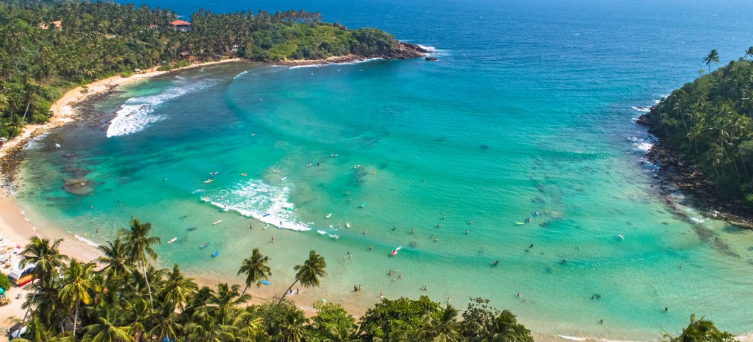 Luftaufnahme einer tropischen Bucht mit türkisblauem Wasser, feinem Sandstrand und vielen Palmen. Zahlreiche Menschen baden oder paddeln auf Surfboards im flachen Wasser. Die Bucht ist von dichter, grüner Vegetation und Felsen umgeben. Im Hintergrund erst