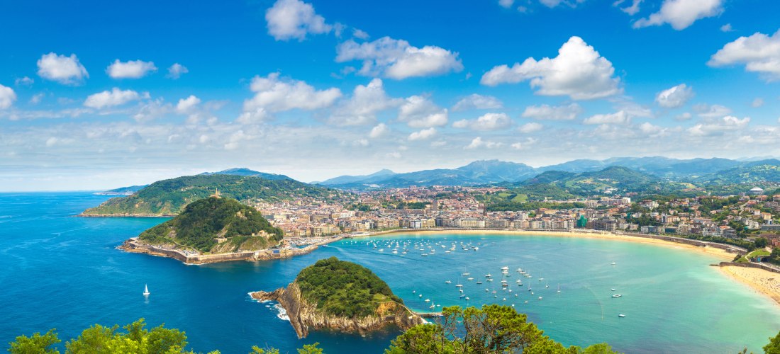 Panoramablick auf San Sebastián mit der muschelförmigen Bucht La Concha, türkisfarbenem Meer, vielen Booten und grünen Hügeln im Hintergrund.