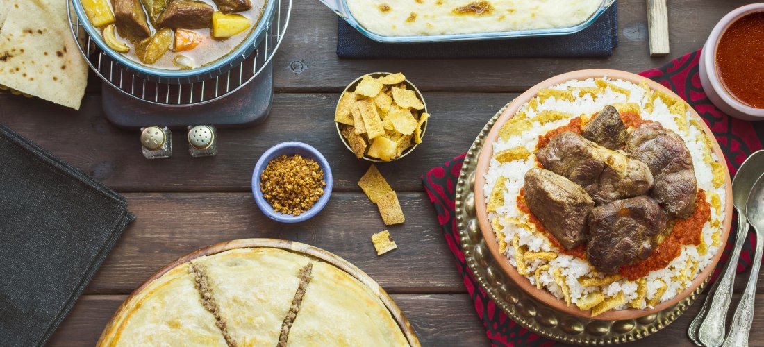 Reich gedeckter Holztisch mit verschiedenen traditionellen Gerichten wie geschmortem Fleisch mit Reis, Fladenbrot, Eintopf mit Gemüse, gebackener Teigkuchen mit Füllung und Beilagen wie Salat, Oliven, Croutons und Saucen.