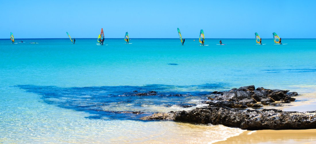 Türkisfarbenes Meer mit mehreren Windsurfern, die auf dem Wasser segeln. Im Vordergrund goldener Sandstrand.