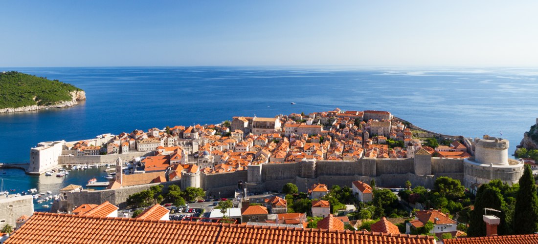 Panoramablick auf die Altstadt von Dubrovnik mit roten Ziegeldächern, die vollständig von einer Stadtmauer umgeben ist. Im Vordergrund das tiefblaue Meer und im Hintergrund eine bewaldete Insel.
