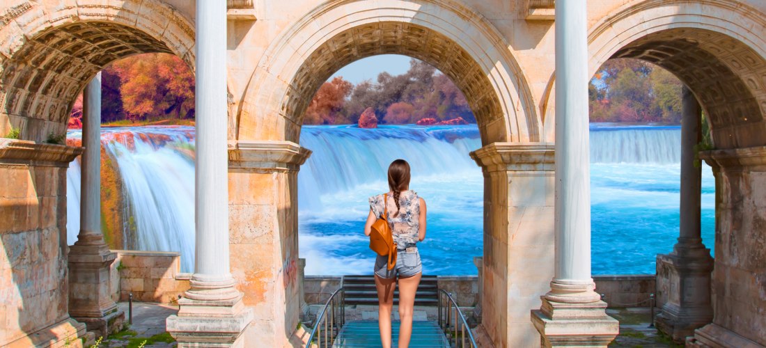 Eine Frau steht auf einem Glassteg zwischen antiken Steinbögen und blickt auf einen breiten Wasserfall mit türkisblauem Wasser. Im Hintergrund sind bunte Bäume und ein sonniger Himmel zu sehen.