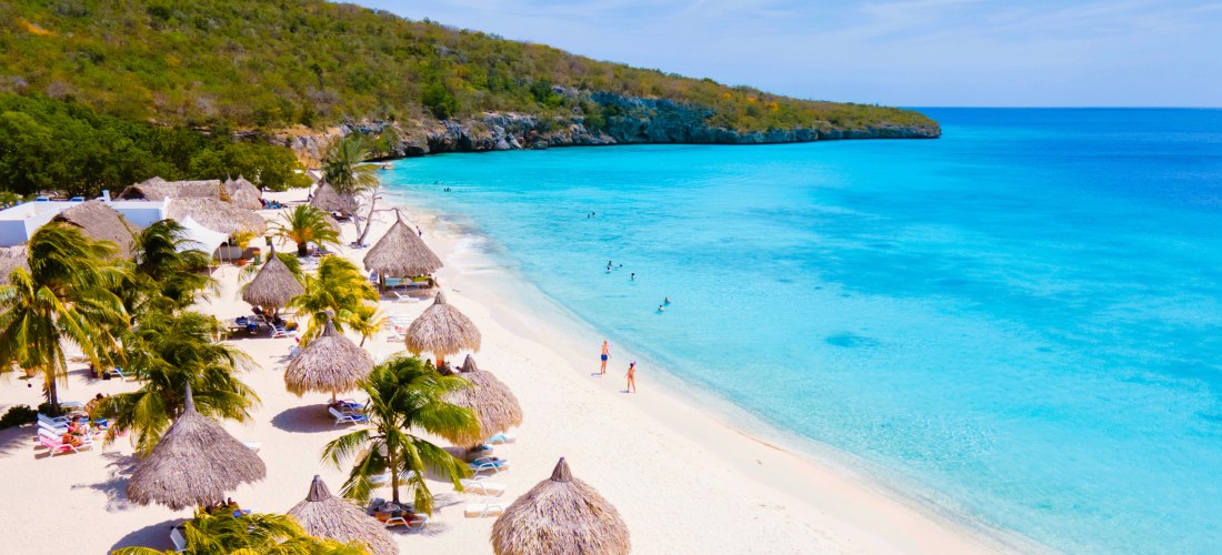 Traumhafter weißer Sandstrand auf Curaçao mit Palmen und strohgedeckten Sonnenschirmen. Türkisblaues, klares Meer mit einigen badenden Menschen. Im Hintergrund eine bewaldete Felsküste unter blauem Himmel mit leichten Wolken