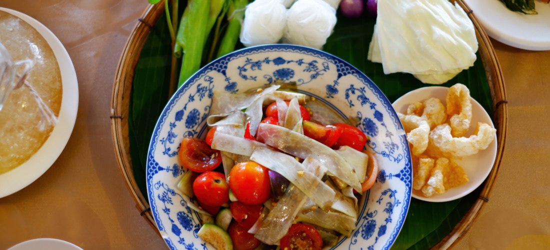 Thailändisches Gericht mit Som Tam (Papayasalat), frischem Gemüse, Reispapier, Reisnudeln, kleinen Auberginen, Weißkohl und knusprigen Schweineschwarten, serviert auf einem runden Bambustablett mit Bananenblatt
