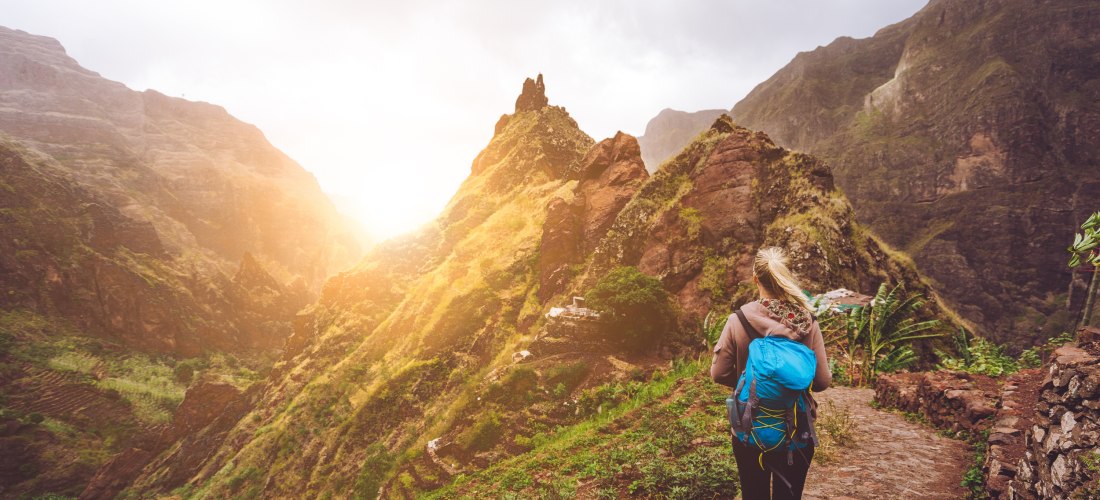 Eine Wanderin mit blauem Rucksack steht auf einem schmalen, gepflasterten Bergweg auf den Kapverden und blickt in ein tiefes, grünes Tal. Um sie herum ragen steile, teils bewachsene Felswände in die Höhe. Die Sonne scheint zwischen den Bergen hervor und t