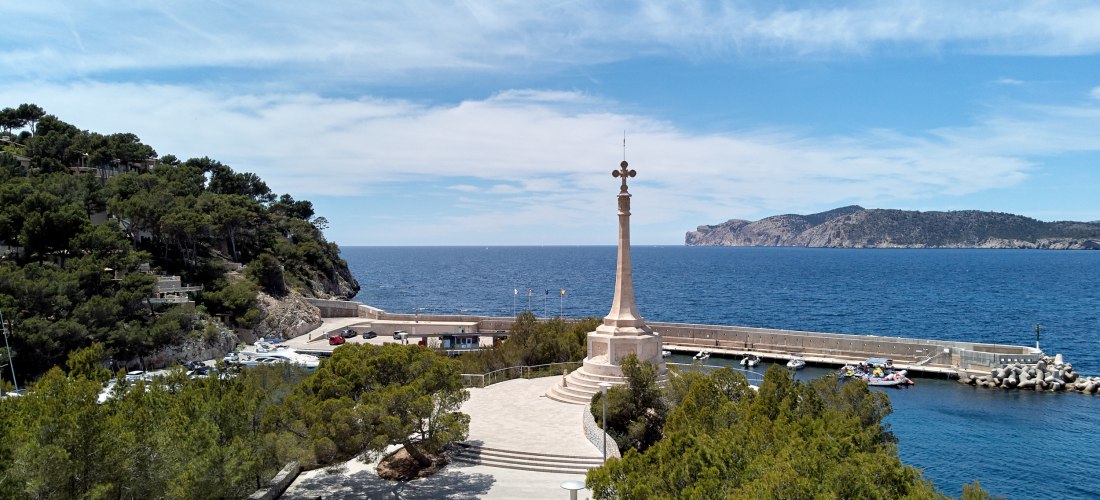 Eine weitläufige Promenade mit einem steinernen Denkmal, das eine hohe Säule mit einem Kreuz trägt. Im Hintergrund das tiefblaue Meer mit einem langen Steg, kleinen Booten und einer felsigen Küste. Die Szene ist von grünem Baumbewuchs gesäumt und unter ei