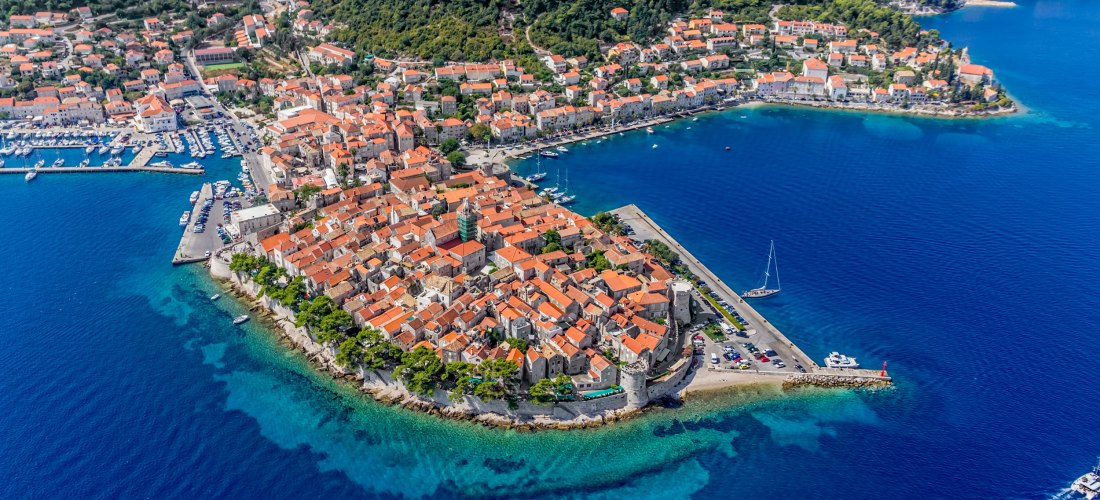 Luftaufnahme der Altstadt von Korčula in Kroatien. Die historische Stadt liegt auf einer Halbinsel und ist von türkisblauem Wasser umgeben. Die roten Ziegeldächer der Häuser und die gut erhaltene Stadtmauer verleihen dem Ort ein mediterranes Flair. Segelb