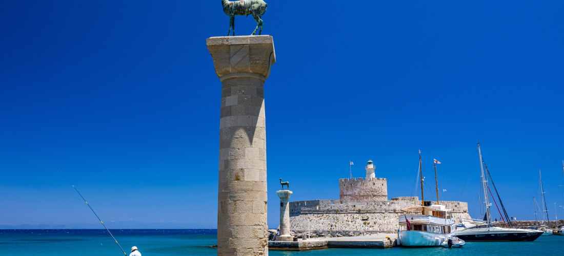 Der Mandraki-Hafen auf Rhodos: Im Vordergrund eine antike Säule mit einer Bronzestatue eines Hirsches, Symbol der Insel. Ein Angler sitzt am Rand des Kais. Im Hintergrund liegt die mittelalterliche Festung Agios Nikolaos, umgeben von Segelbooten und türki