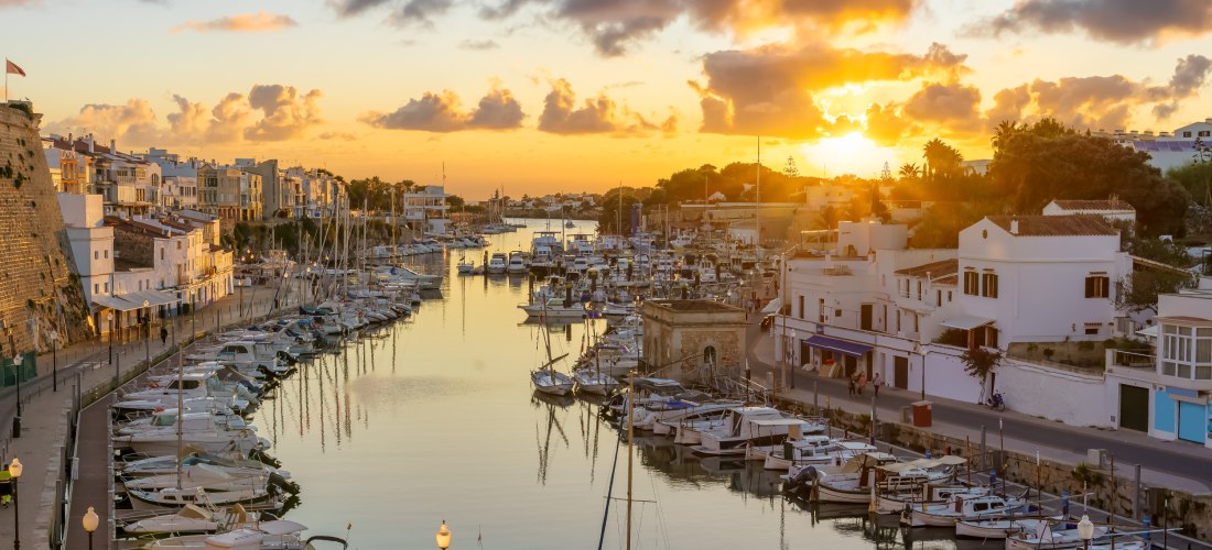 Hafen von Ciutadella de Menorca bei Sonnenuntergang. Zahlreiche Boote und Yachten liegen im Wasser, umgeben von weißen Häusern und alten Stadtmauern.
