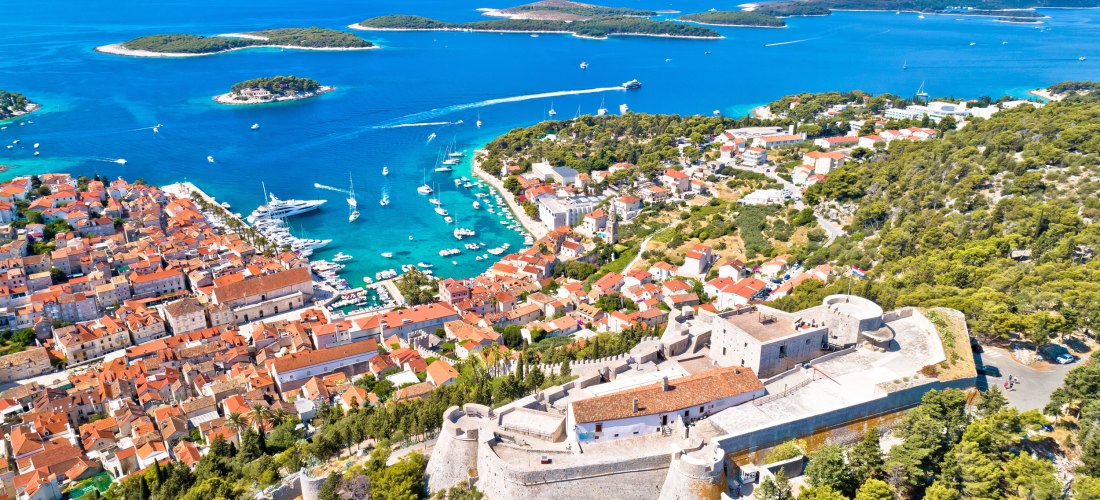 Kroatien - Hvar Stadt Luftaufnahme von Hvar-Stadt auf der gleichnamigen Insel in Kroatien. Die Altstadt mit ihren roten Ziegeldächern zieht sich entlang der Bucht, in der zahlreiche weiße Boote und Yachten liegen. Im Vordergrund thront die historische Festung Fortica auf einem