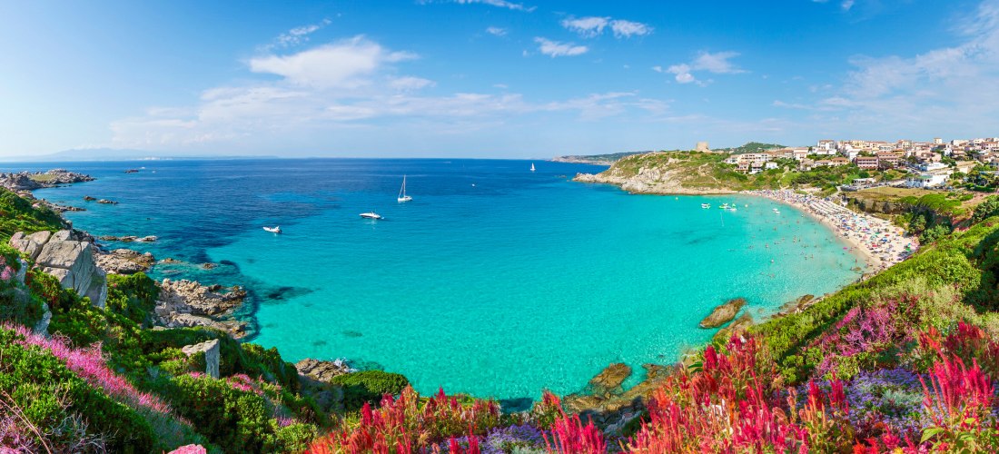 Türkisfarbene Bucht mit Sandstrand, felsiger Küste und einer Kleinstadt am Hang. Im Vordergrund blühende Pflanzen in kräftigen Farben.