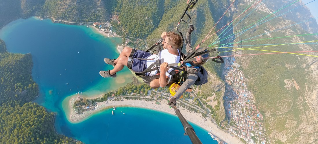 Paragliding über der türkisblauen Lagune von Ölüdeniz in der Türkei. Zwei Personen gleiten im Tandemflug durch die Luft, unter ihnen die grüne Küstenlandschaft, das Meer und kleine Boote.